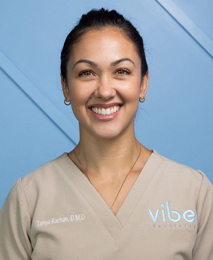 Female dental professional smiling at camera wearing Vibe Dentistry scrubs against blue background.