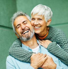 Smiling mature couple embracing outdoors against green background, representing dental care and wellness.