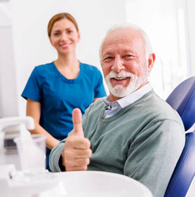 Elderly man giving thumbs up in dental chair with hygienist in background at modern dental office.