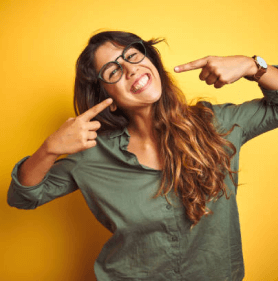 Woman pointing at her smile wearing glasses against yellow background, promoting dental care.