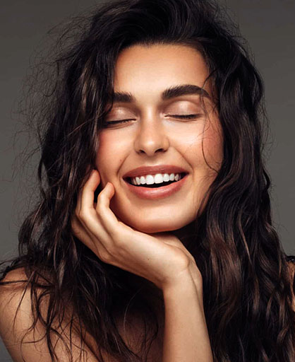 Woman with dark wavy hair smiling brightly, hand on face, against neutral gray background.